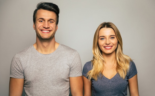 Photo en gros plan d'un beau couple, qui pose dans des t-shirts gris, debout les bras baissés, regardant dans la caméra et souriant