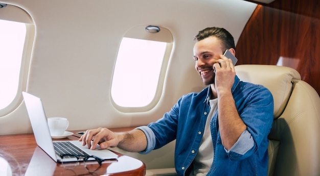 Photo en gros plan d'un bel homme dans une tenue décontractée, qui sourit, tout en tapant quelque chose sur son ordinateur portable et tenant un smartphone dans sa main gauche, volant en première classe d'avion.