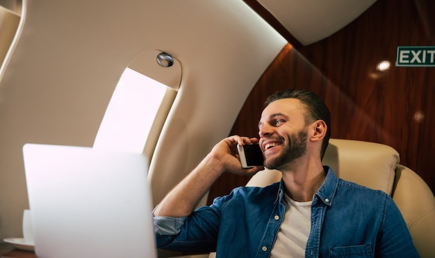 Photo en gros plan d'un bel homme dans une tenue décontractée, qui sourit, tout en tapant quelque chose sur son ordinateur portable et tenant un smartphone dans sa main gauche, volant en première classe d'avion.