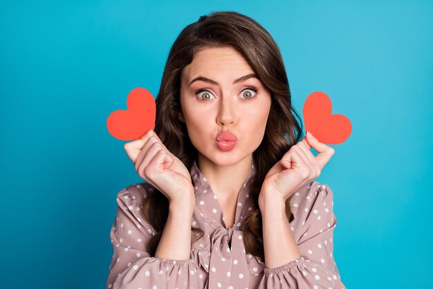 Photo en gros plan de la belle charmante dame drôle tenir deux petites cartes de coeur rouge humeur romantique envoyant des baisers aériens petit ami porter une robe en pointillés fond de couleur bleu isolé