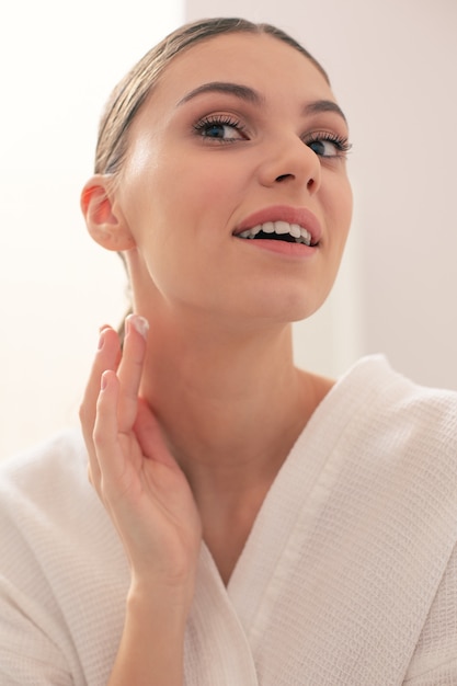 Photo en gros plan de la belle jeune femme debout seule et souriante tout en mettant une petite quantité de crème pour le visage sur son cou