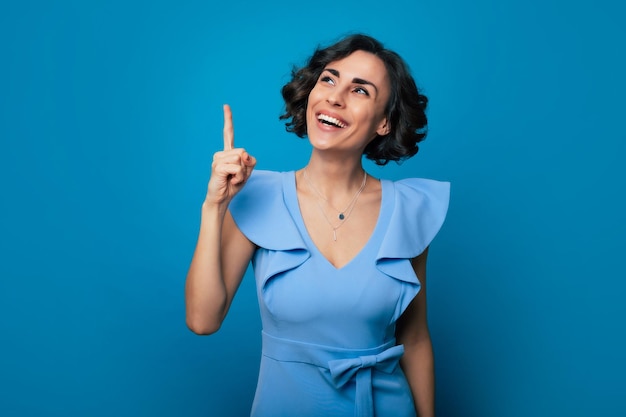 Photo en gros plan d'une belle jeune femme heureuse dans une robe bleue, qui pointe avec son index et isolée sur fond bleu