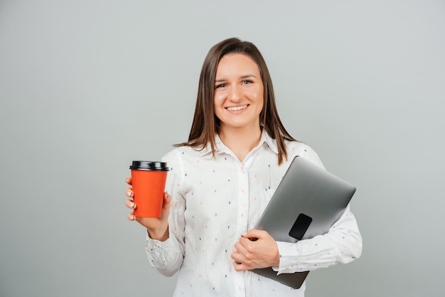 Photo en gros plan d'une charmante jeune étudiante tenant une tasse de café en papier rouge et tenant un ordinateur portable