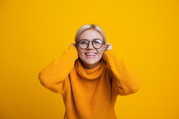 La photo en gros plan d'une dame de race blanche aux cheveux blonds et des lunettes touchant ses cheveux et souriant à l'avant sur un fond jaune