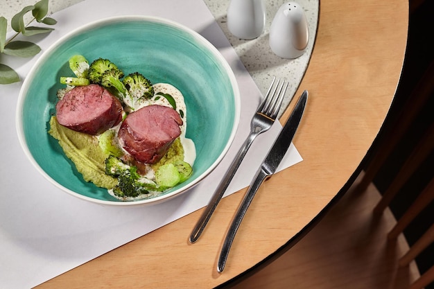 Photo en gros plan d'une délicieuse viande rôtie avec du brocoli et des courgettes assaisonnées de sauce Servi sur une assiette creuse bleue Couverts Concept de cuisine de restaurant Vue de dessus