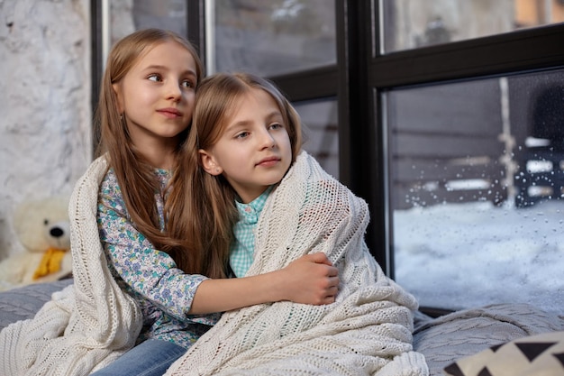 Photo en gros plan de deux belles petites sœurs caucasiennes assises sur le rebord de la fenêtre, couvertes d'une couverture. Ils se sont figés et ont essayé de se réchauffer, utilisant une couverture se serrant les uns contre les autres