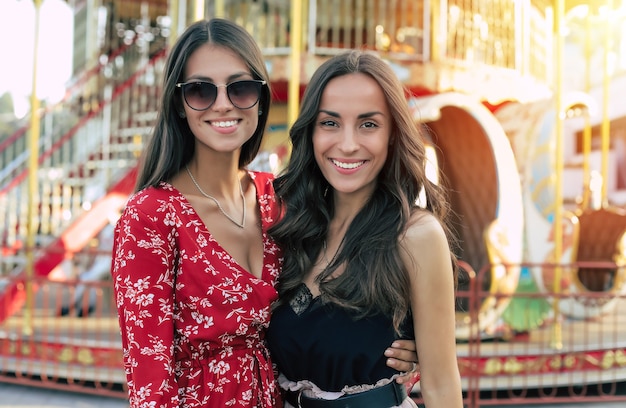 Photo en gros plan de deux jeunes amies séduisantes en robes de fête, posant devant le carrousel, regardant la caméra et souriant de joie