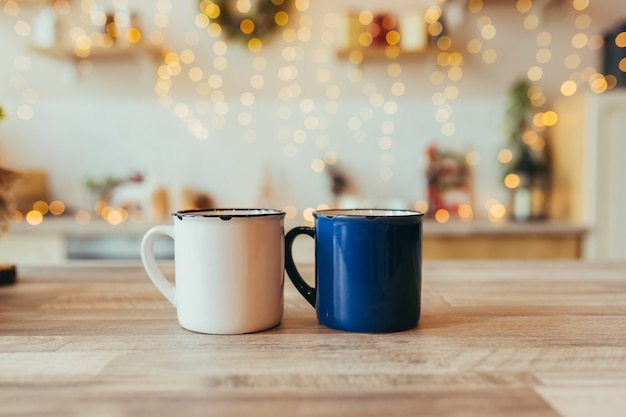 Photo en gros plan de deux tasses de Noël, sur fond de nouvel an, arbre de Noël et lumières colorées, dans la cuisine sur la table