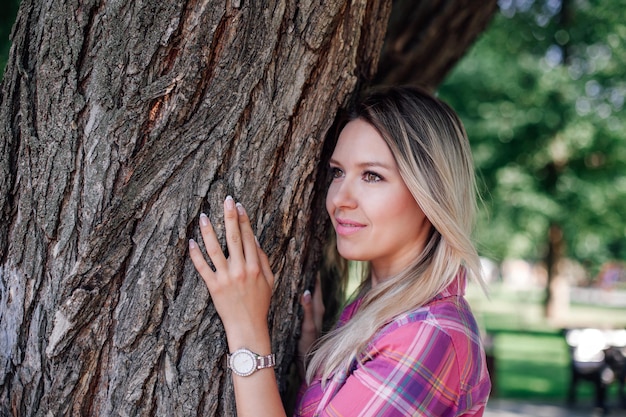 Photo gros plan d'une femme blonde brillante adulte avec le visage regardant loin debout dans un parc s'appuyant sur un chêne...