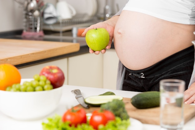 Photo gros plan d'une femme enceinte posant avec une pomme verte dans la cuisine