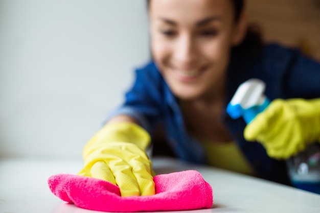 Photo en gros plan d'une femme portant des gants de protection souriante et essuyant la poussière à l'aide d'un vaporisateur et d'un plumeau tout en nettoyant sa maison