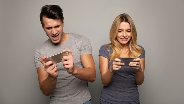 Photo en gros plan d'une fille blonde et de son beau petit ami, qui jouent à des jeux mobiles sur leurs smartphones avec des expressions faciales concentrées.