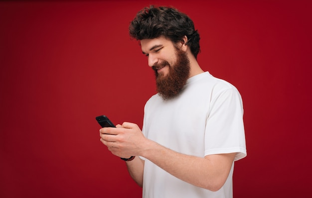 La photo en gros plan d'un homme barbu à l'aide d'un mobile, vêtu d'un t-shirt blanc sur un mur rouge