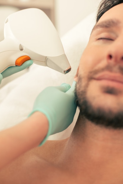 Photo photo en gros plan de l'homme barbu avec les yeux fermés et les mains dans des gants en caoutchouc effectuant l'épilation au laser et touchant l'oreille de l'homme