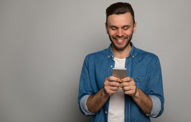 Photo en gros plan d'un homme gai dans une veste en jean, qui sourit, tout en tenant un smartphone dans ses mains.