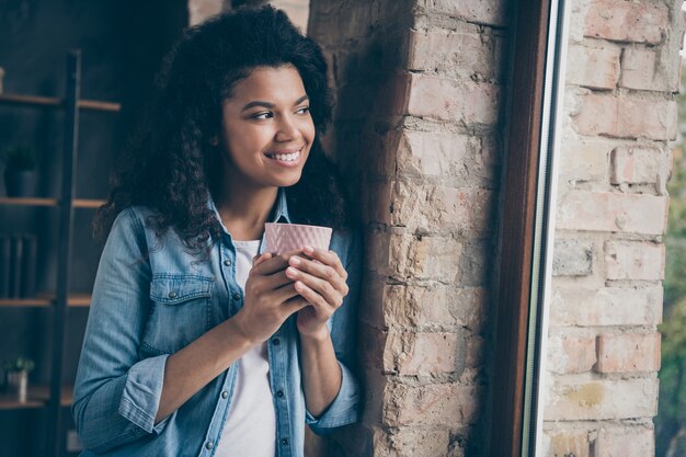 Photo gros plan de l'incroyable peau foncée curly lady holding hot coffee drink à la fenêtre penchée mur de brique porter des appartements modernes tenue en denim décontracté à l'intérieur