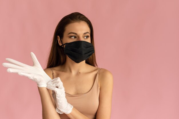 La Photo En Gros Plan D'une Jeune Femme Dans Un Masque Médical Met Des Gants Stériles Chirurgicaux Protecteurs Sur Son Bras Sur Fond Rose, Protection Contre Le Coronavirus.