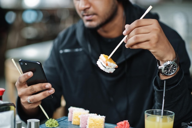 Photo en gros plan d'un jeune homme asiatique décontracté et élégant au café mangeant des sushis et regardant un téléphone portable