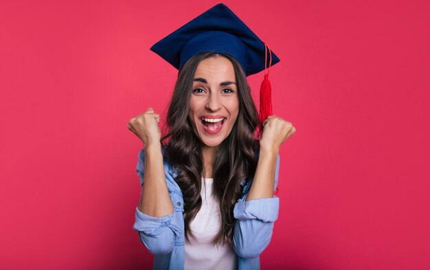 Photo en gros plan d'une jolie étudiante vêtue d'un chemisier et d'un chapeau académique carré, qui est ravie et crie de bonheur pour son succès