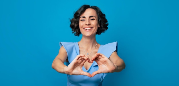 Photo en gros plan d'une jolie femme joyeuse dans une longue robe bleue qui regarde dans l'appareil photo et souriant tout en montrant un coeur avec ses mains isolées sur fond bleu