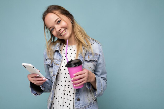 Photo en gros plan d'une jolie femme magnifique en veste en jean souriante et tenant un smartphone en buvant