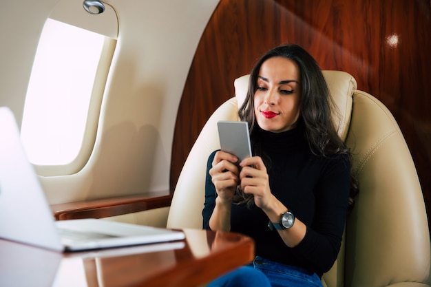 Photo en gros plan d'une jolie fille vêtue d'une tenue décontractée, qui lit quelque chose sur son smartphone, tout en prenant un vol confortable à bord d'un avion de première classe.