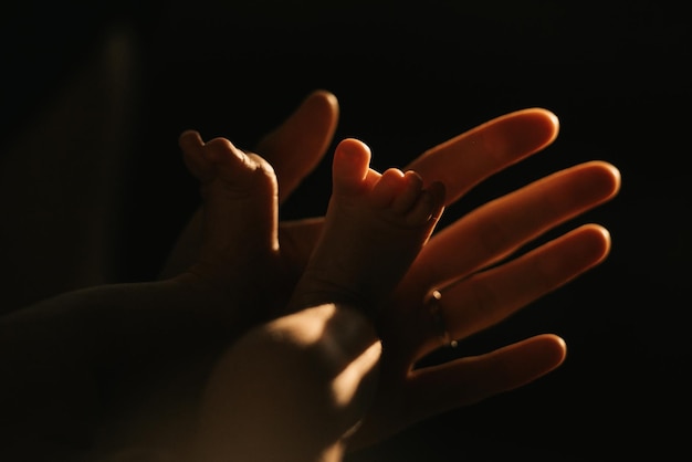 Une photo en gros plan de la main douce de la mère qui tient les pieds de son nouveau-né. Les jambes du nourrisson au soleil le soir.