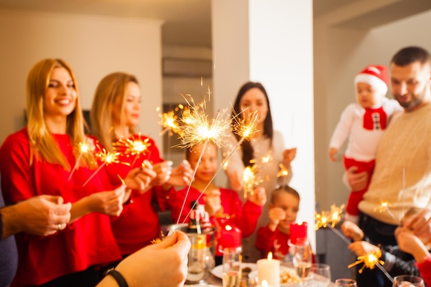 Photo en gros plan d'une main de femme tenant des étincelles allumées, groupe d'hommes et de femmes souriants heureux avec des enfants sur un fond, tenant des étincelles, estompées. Célébration de Noël