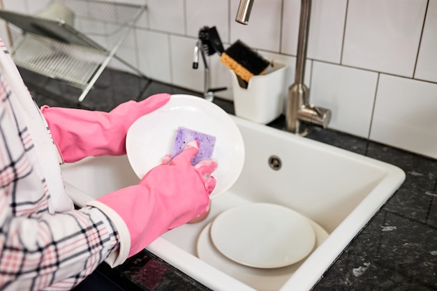 Photo en gros plan de mains féminines lavant une assiette blanche avec une éponge et de la mousse dans un évier de cuisine. Concentrez-vous sur les mains dans des gants en caoutchouc rose