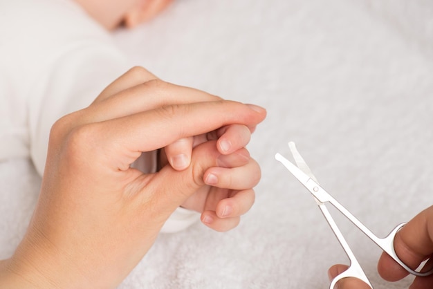 Photo en gros plan des mains de la mère tenant les doigts du nouveau-né et les ciseaux à ongles pour bébé sur fond textile blanc isolé