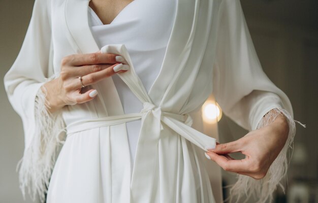 Photo en gros plan d'une mariée en robe de chambre en soie tenant une robe de mariée devant la fenêtre Les préparatifs de la mariée du matin du mariage