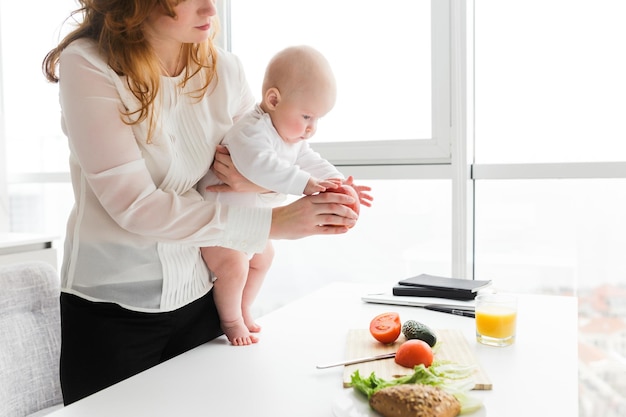 Photo en gros plan d'une mère debout et tenant son mignon petit bébé tout en lui montrant une grosse pomme rouge dans la cuisine