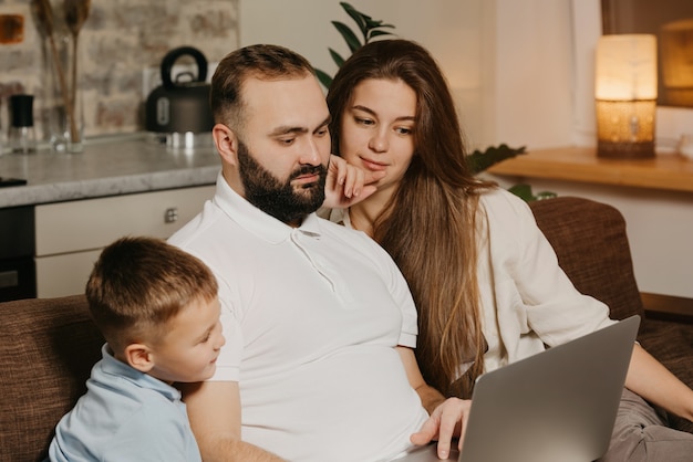 Une photo en gros plan d'un père avec une barbe qui montre ses réalisations au travail sur un ordinateur portable à son fils et à sa femme pensive à la maison. Papa travaille à distance sur un ordinateur entre proches le soir