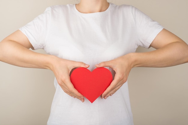 Photo en gros plan recadrée d'une fille en t-shirt blanc tenant un coeur en papier rouge près du haut de l'abdomen sur fond gris isolé