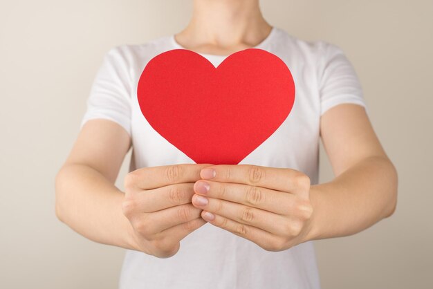 Photo en gros plan recadrée d'une jeune femme en t-shirt blanc donnant un coeur de papier rouge sur fond gris isolé avec un espace vide