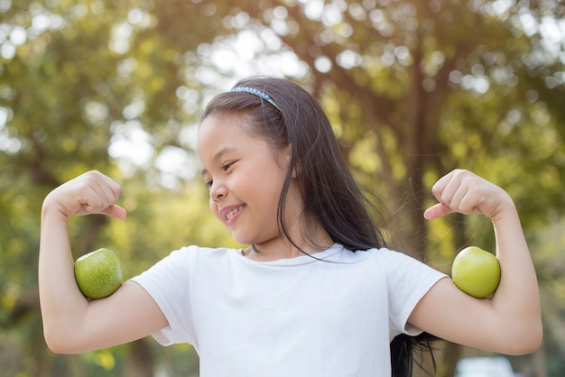 Photo heureuse petite fille asiatique enfant debout avec un grand sourire. fille avec pomme verte montrant les biceps. Nature bio verte saine et fraîche avec feuillage flou abstrait et lumière du soleil d'été