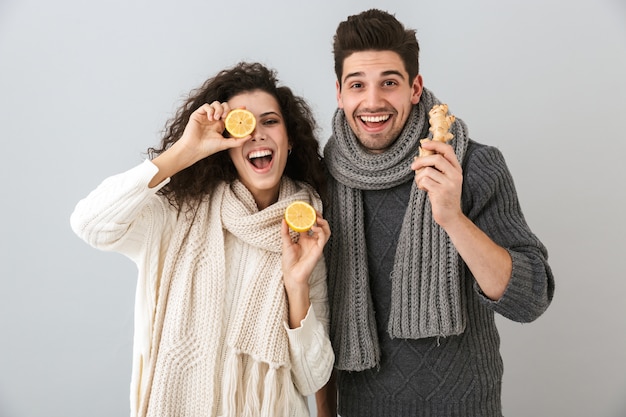 Photo d'heureux couple homme et femme portant des écharpes tenant des citrons et du gingembre, isolé sur mur gris
