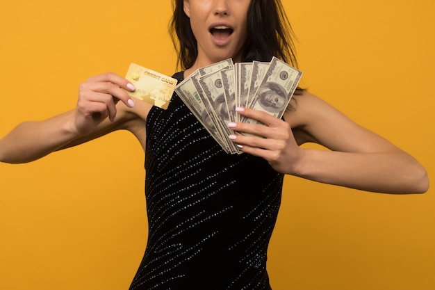 Photo de heureux heureux jeune femme posant isolé sur fond de mur jaune tenant de l'argent et une carte de crédit ou de dette.