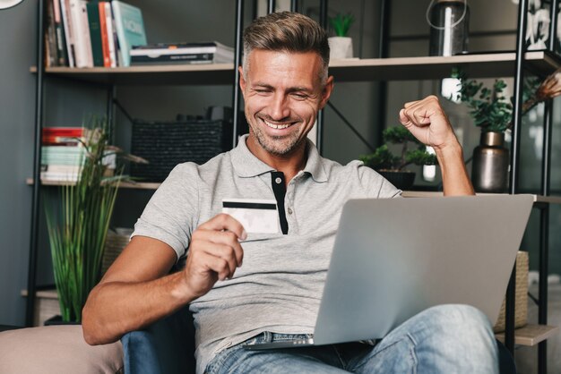 Photo d'un homme adulte joyeux en vêtements décontractés tenant une carte de crédit et un ordinateur portable assis sur un fauteuil dans un appartement