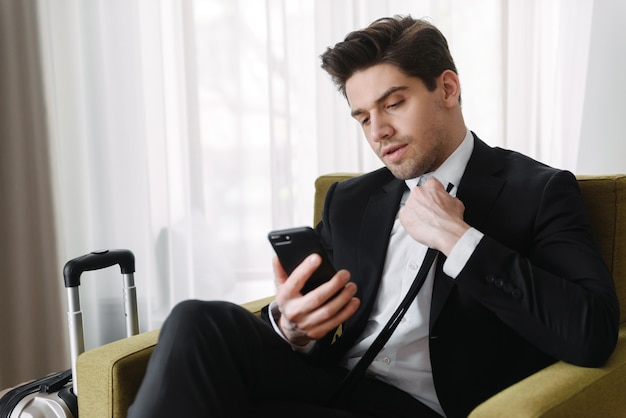 Photo d'un homme d'affaires brune maussade portant un costume noir en train de taper sur un téléphone portable alors qu'il était assis sur un fauteuil dans un appartement de l'hôtel