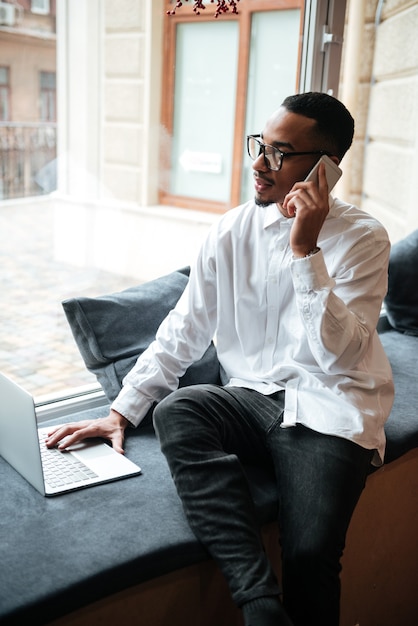 Photo d'un homme d'affaires séduisant portant des lunettes assis près de la fenêtre et tapant par ordinateur portable tout en parlant par téléphone