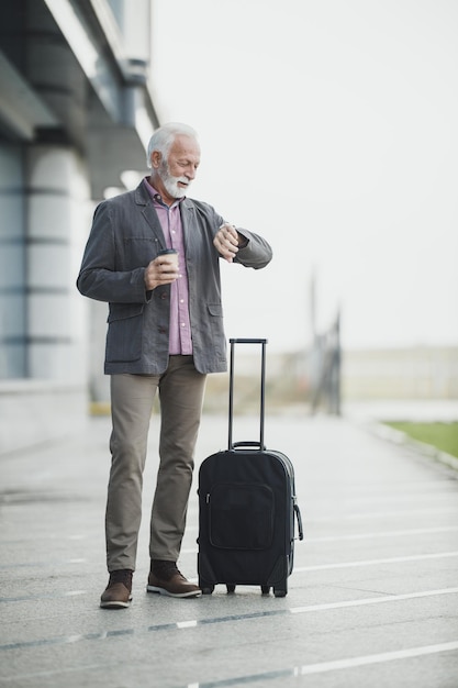 Photo d'un homme d'affaires senior debout avec ses bagages et regardant une montre à main à l'extérieur de l'aéroport.