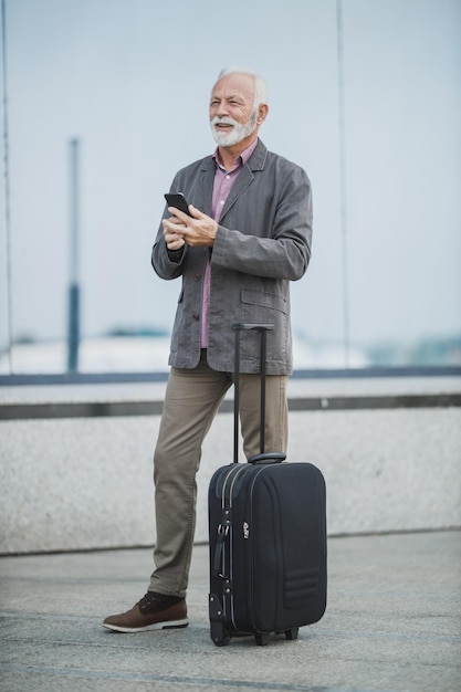 Photo d'un homme d'affaires senior marchant avec ses bagages et utilisant un smartphone à l'extérieur de l'aéroport.