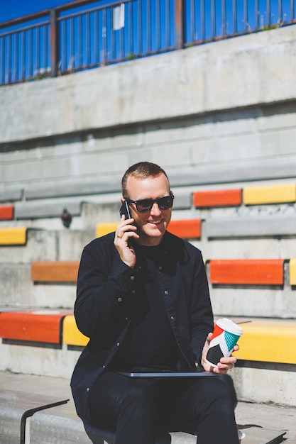 Photo d'un homme d'affaires souriant en costume tenant un téléphone portable tout en se tenant à l'extérieur près d'un bâtiment avec du café à emporter