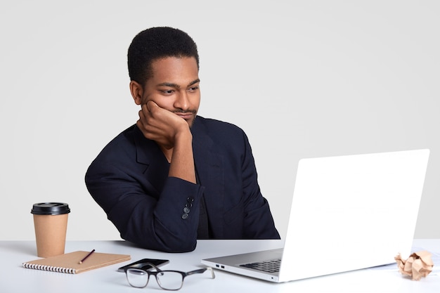 Photo d'un homme afro-américain à la peau sombre qui garde la main sur la joue, regarde attentivement le didacticiel vidéo, se prépare pour une réunion d'affaires, a un bloc-notes et un stylo sur un bureau blanc, porte un costume formel, pose à l'intérieur