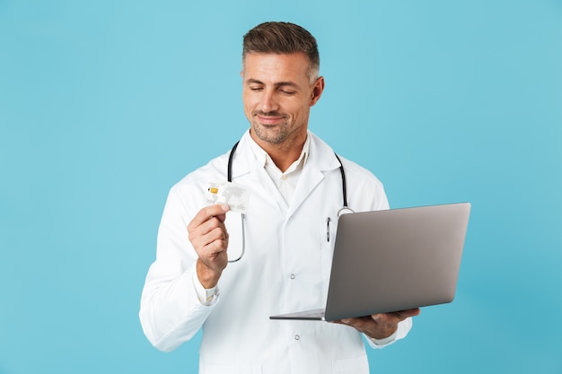 Photo d'un homme d'âge moyen portant une blouse médicale blanche et un stéthoscope tenant un ordinateur portable et une carte de crédit, debout isolé sur un mur bleu