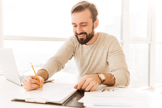 Photo D'un Homme D'âge Mûr Réussi Dans Des Vêtements Décontractés Souriant Et écrivant Des Notes Sur Un Document Papier, Tout En Travaillant Dans La Chambre Avec Fenêtre