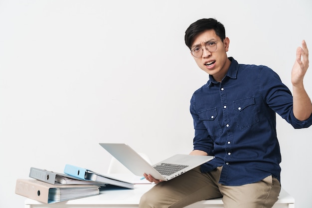 Photo d'un homme asiatique perplexe portant des lunettes assis à table et travaillant sur un ordinateur portable au bureau isolé sur un mur blanc