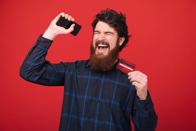 Photo d'un homme barbu, célébrant son premier salaire, les mains levées et les yeux fermés, mur rouge