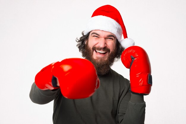 Photo d'un homme barbu excité portant un chapeau de père Noël frappant avec des gants de boxe rouges